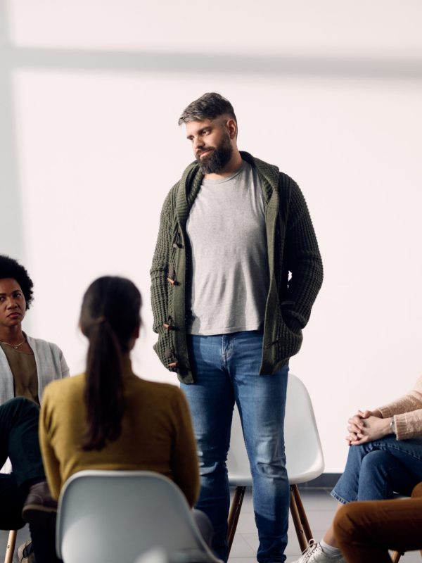 Participants of group therapy sitting in a circle while one of them is standing and talking about his issues during a session. Copy space.